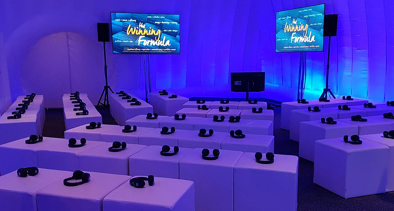 Pic showing the inside of a Created By Air inflatable event structure setup with seating, blue lighting and several TV screens displaying The Winning Formula on the screens
